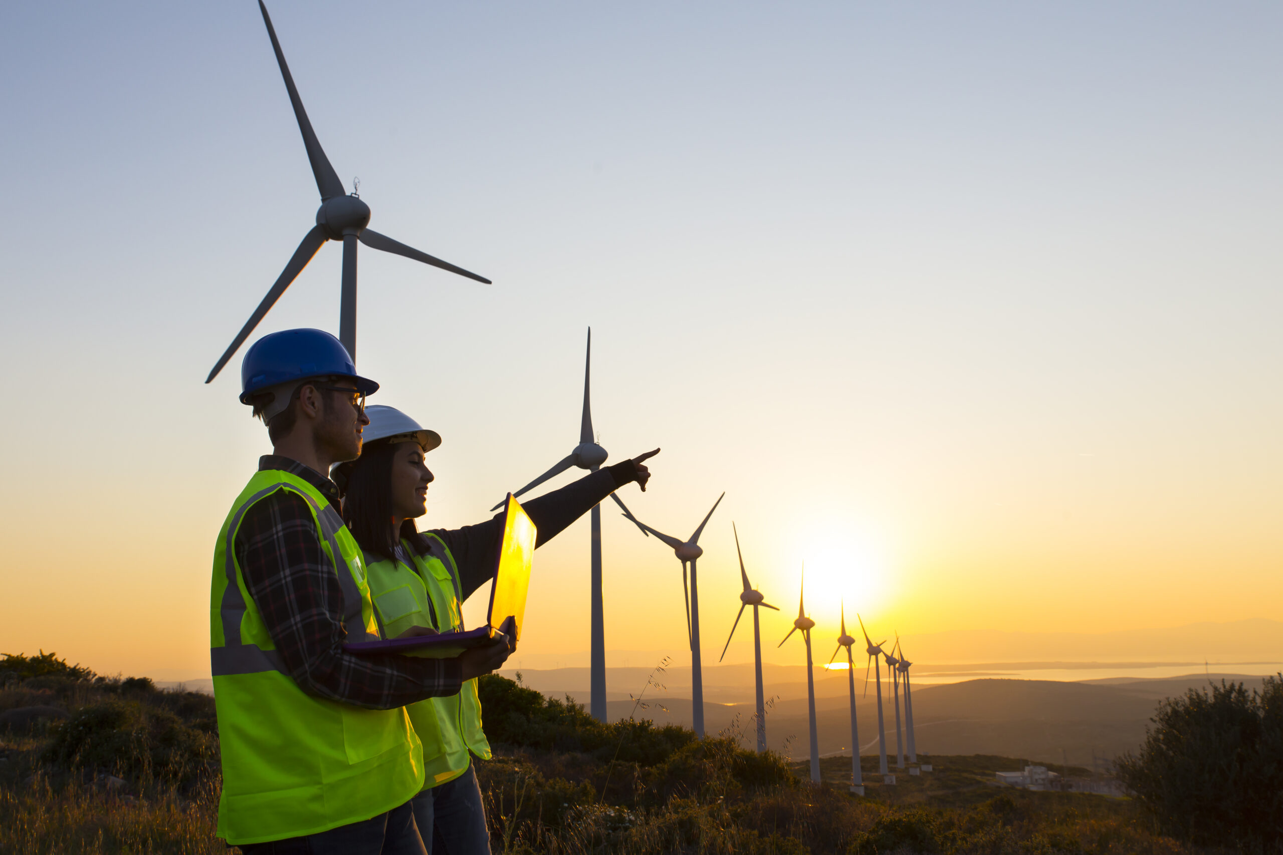 Two men in high vis vests and hard hats pointing and holding a computer at a windmill farm during sunset.