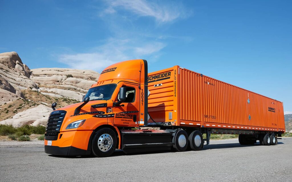 Large orange truck driving on a road with blue skies and a hill behind.